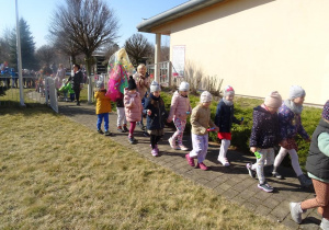 Dzieci maszerują z wiosennymi plakatami, panią Wiosną pod budynkiem przedszkola.